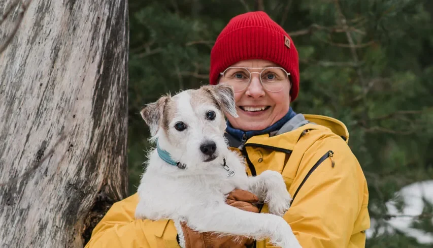 Smiling person in a yellow jacket and red knit cap holding a small white dog in a snowy forest beside a tree.